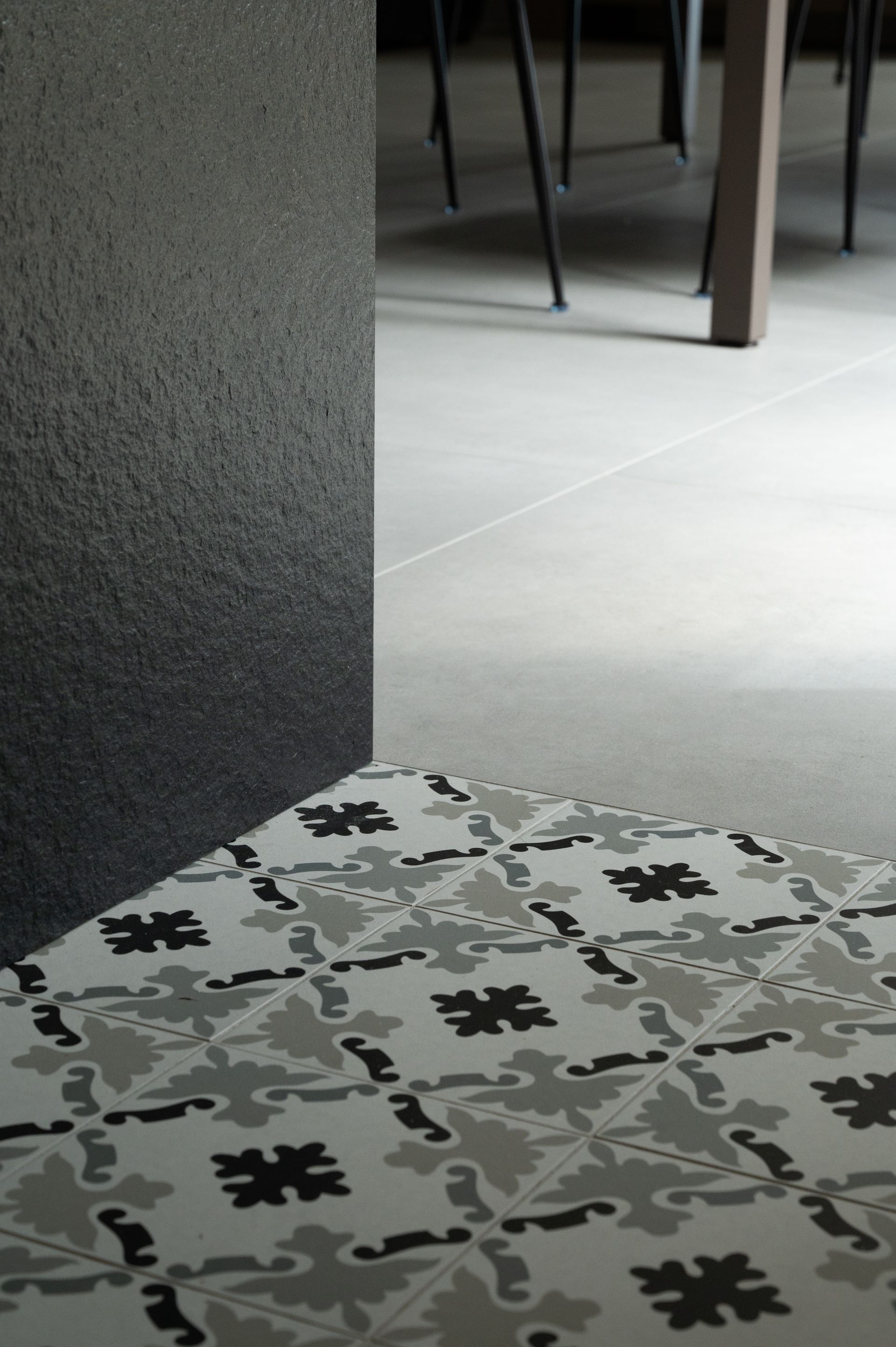 Coin de carrelage à motifs, mur texturé foncé, sol gris clair et pieds de table.
