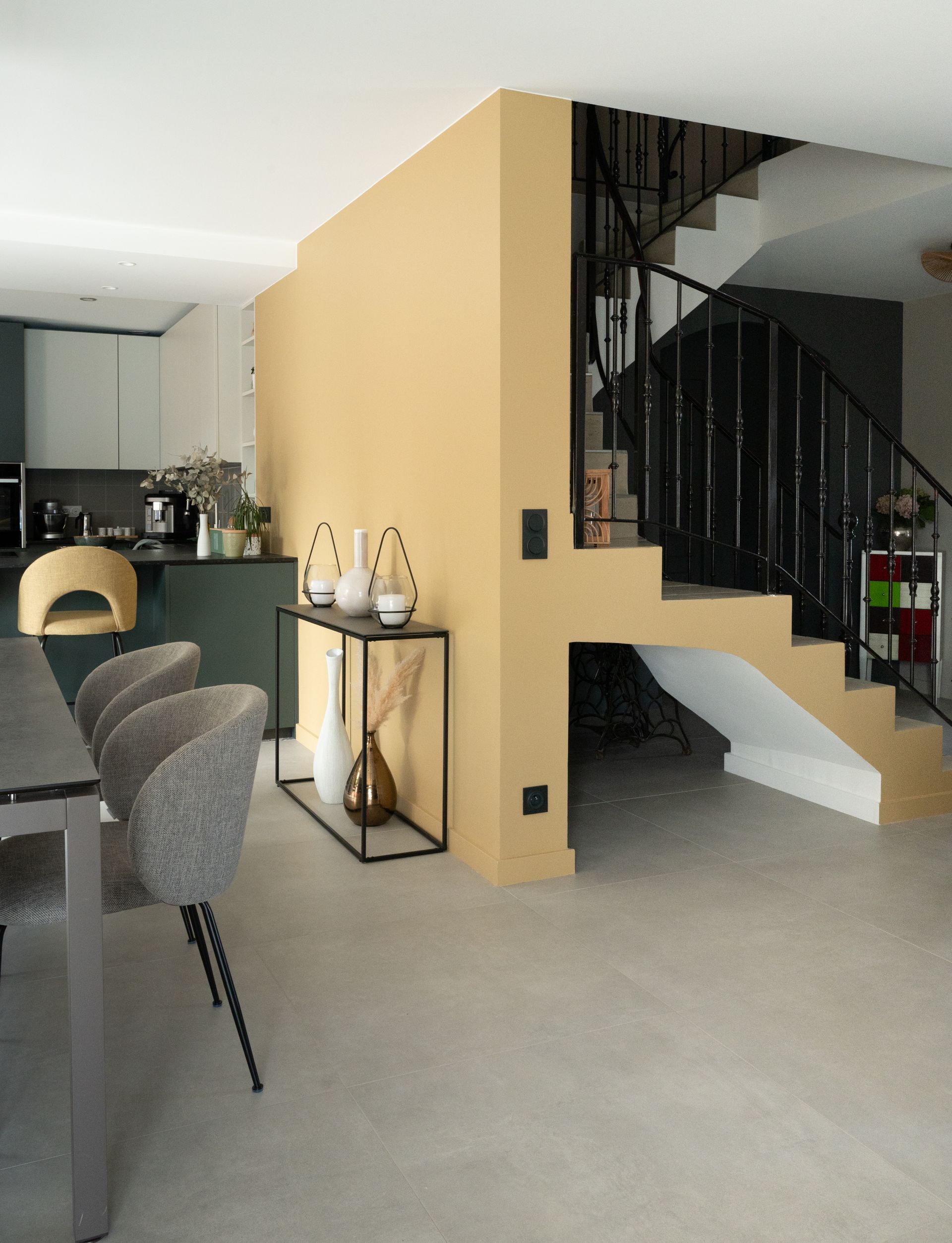 Intérieur moderne avec escalier, coin repas et cuisine. Mur beige, escalier noir, chaises grises.