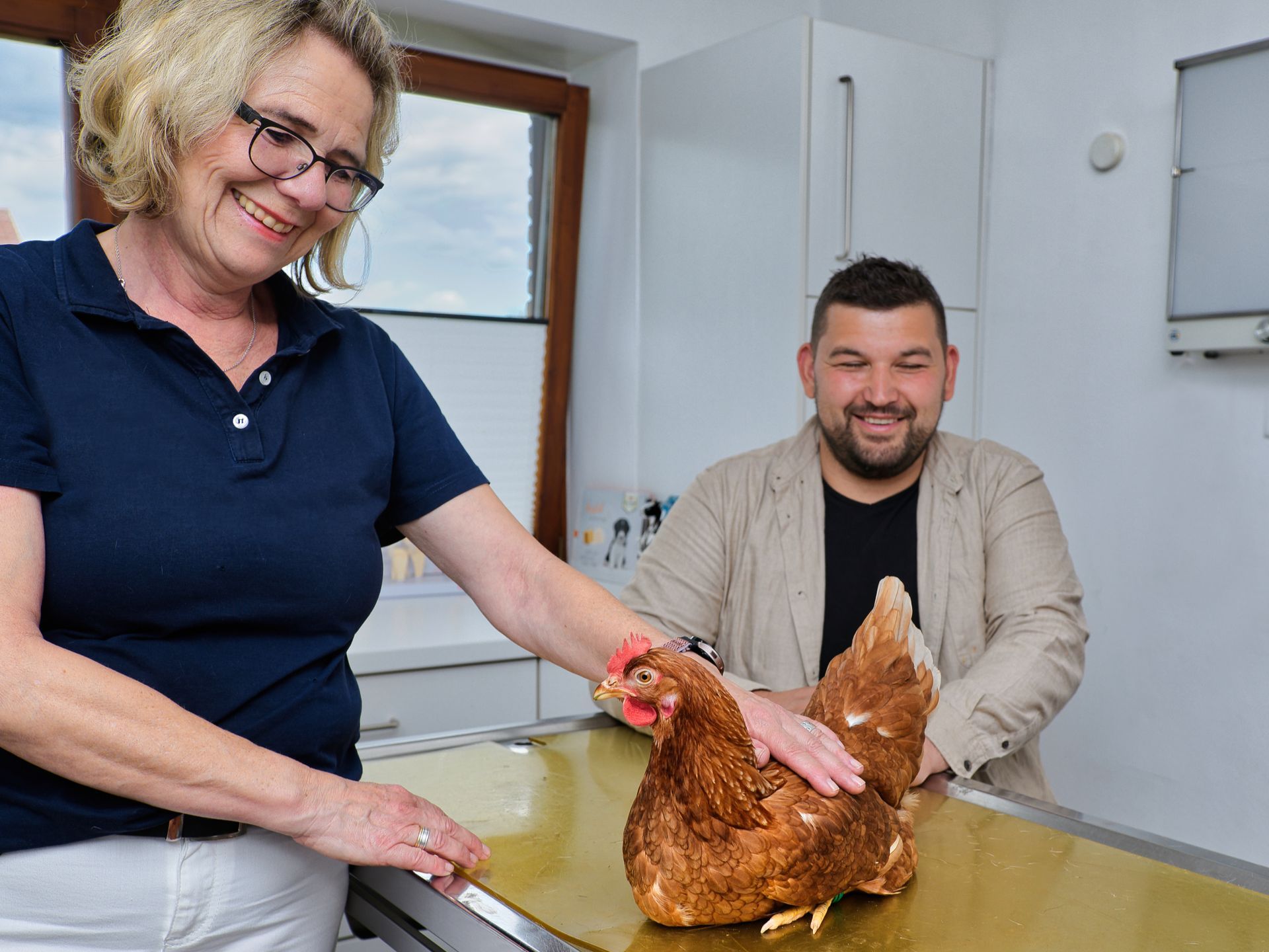 Tierärztin untersucht ein braunes Huhn auf einem Untersuchungstisch; Mann schaut zu. Klinikinneres, lächelnde Gesichter.