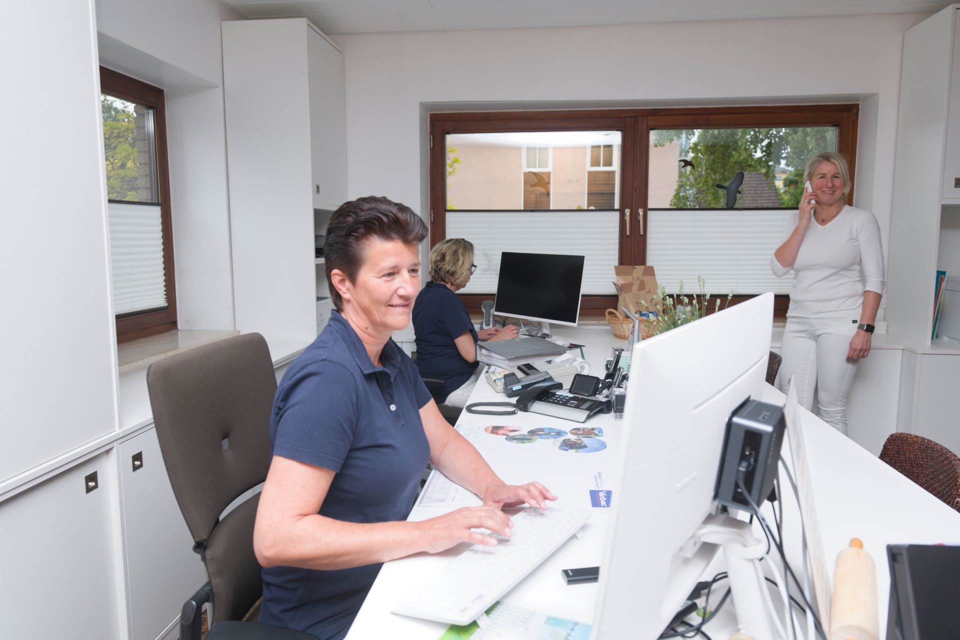 Drei Frauen an Schreibtischen in einem Büro; eine tippt, eine andere arbeitet am Computer und eine telefoniert.