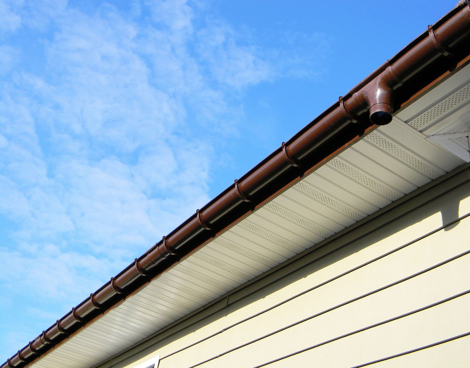 Close-up of a house roof eave with brown gutter and cream siding against a blue sky