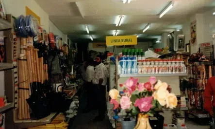 Un estrecho pasillo de tienda con estanterías llenas de mercancía y flores al frente, iluminado con luces fluorescentes.