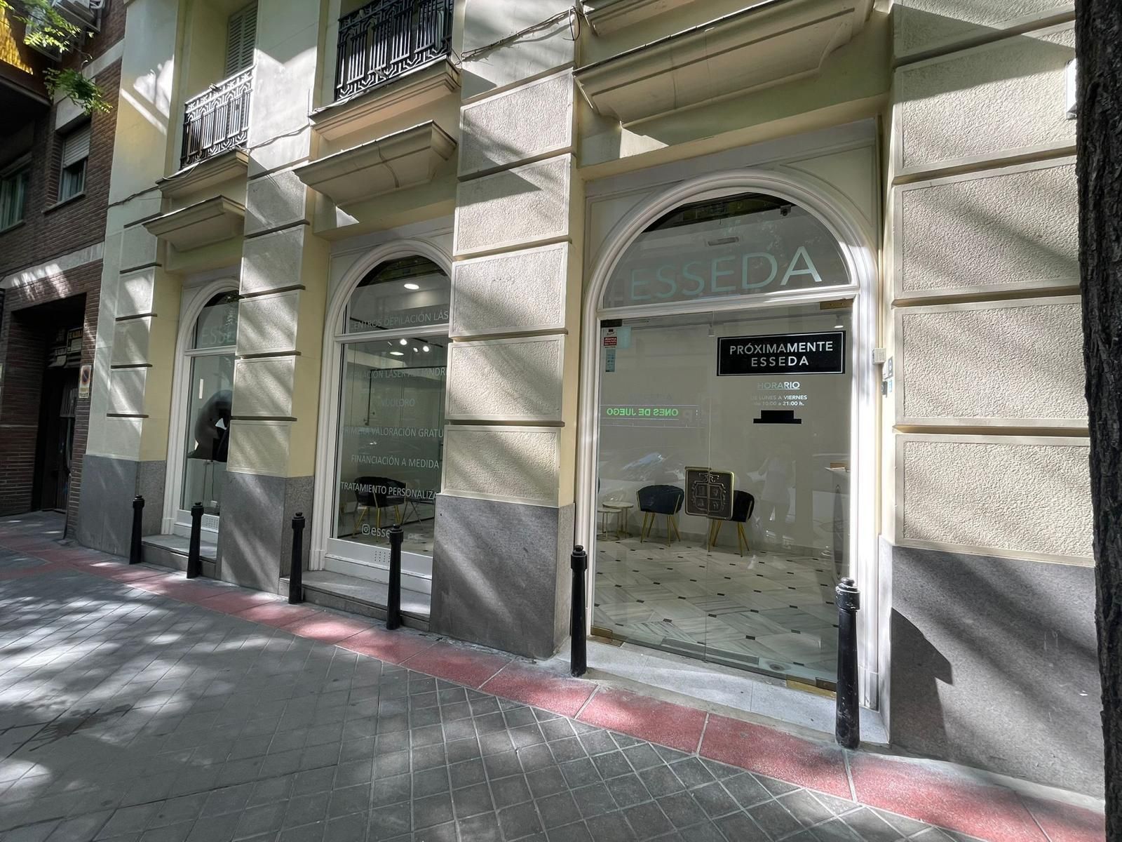 Local comercial con ventanas arqueadas y puertas negras situado en una acera soleada bajo el balcón de un edificio.