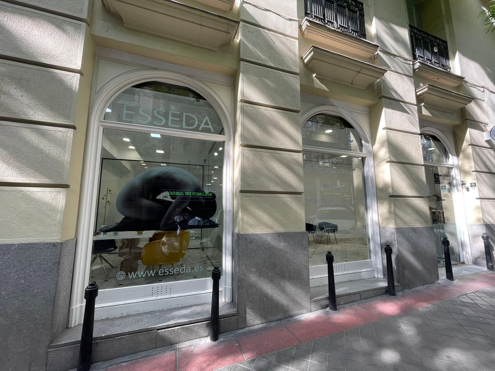 Escaparates a pie de calle en un edificio de piedra clara, con cristales arqueados y reflejos de los coches aparcados.