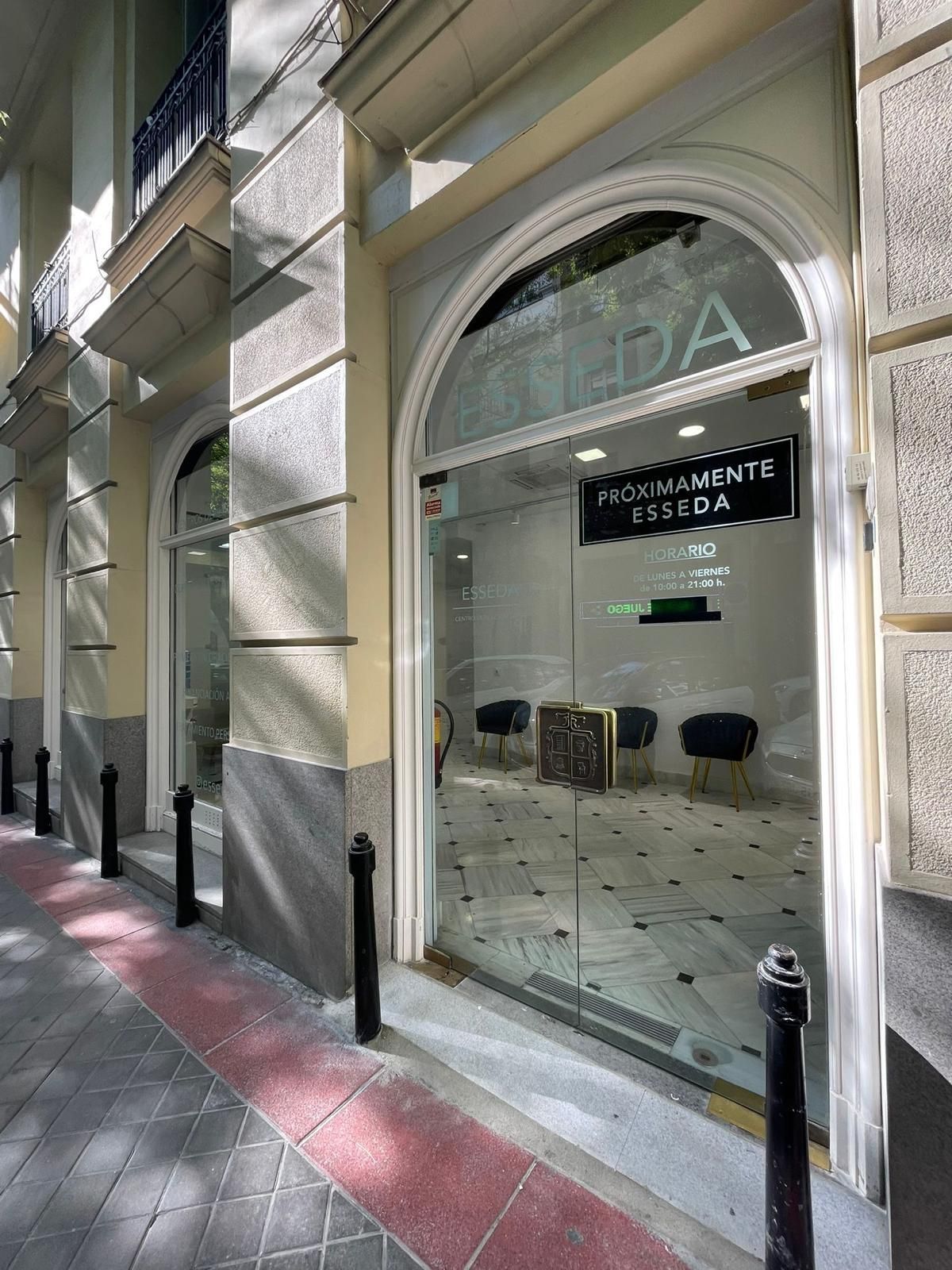 Local comercial con puerta de cristal arqueada, sillas negras en el interior y una acera embaldosada en el exterior.