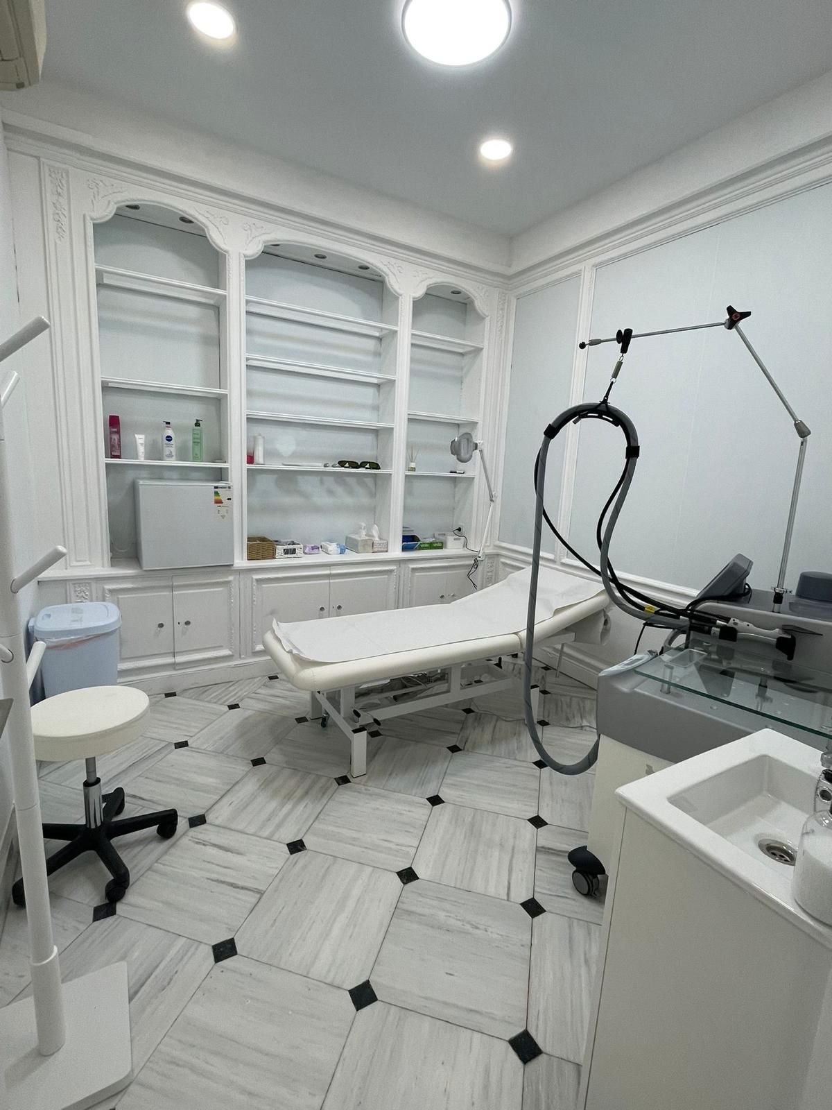 Sala de tratamiento de color blanco brillante con sillón de exploración, armarios, equipo y lavabo en una clínica.