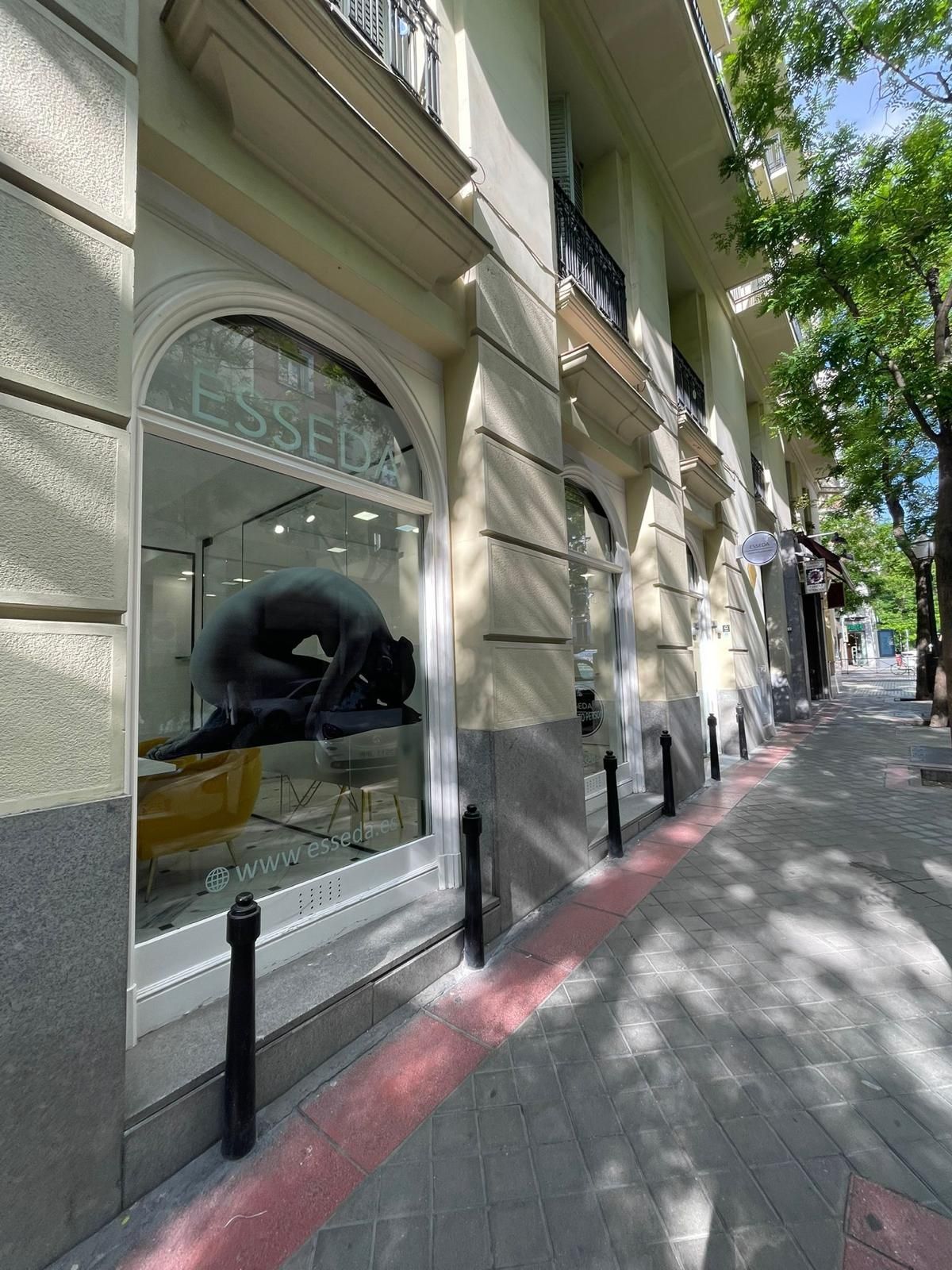 Acera junto a un edificio beige con ventanas arqueadas, sombras y una persona vista a través del escaparate de una tienda.
