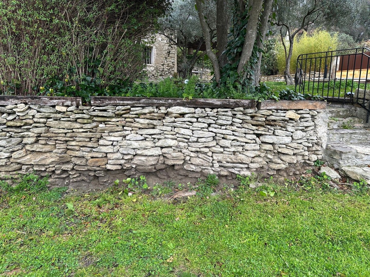 Un mur de pierre se dresse devant une pelouse verdoyante. Derrière le mur se trouvent des arbres et un portail en fer forgé.