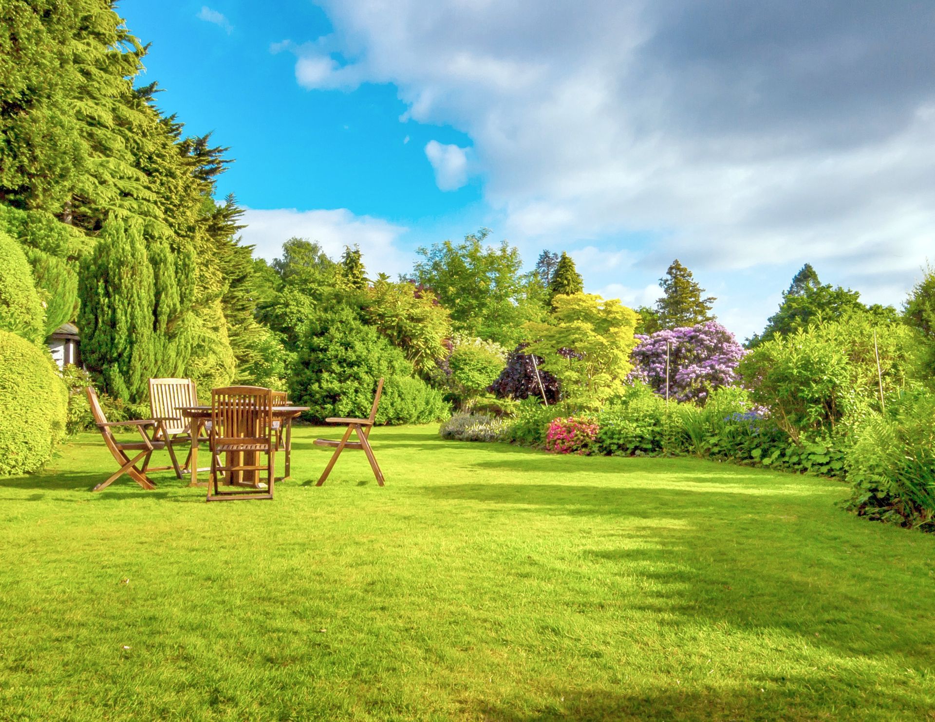 Jardin arrière verdoyant et luxuriant avec mobilier de jardin, entouré d'arbres et de fleurs sous un ciel bleu nuageux.