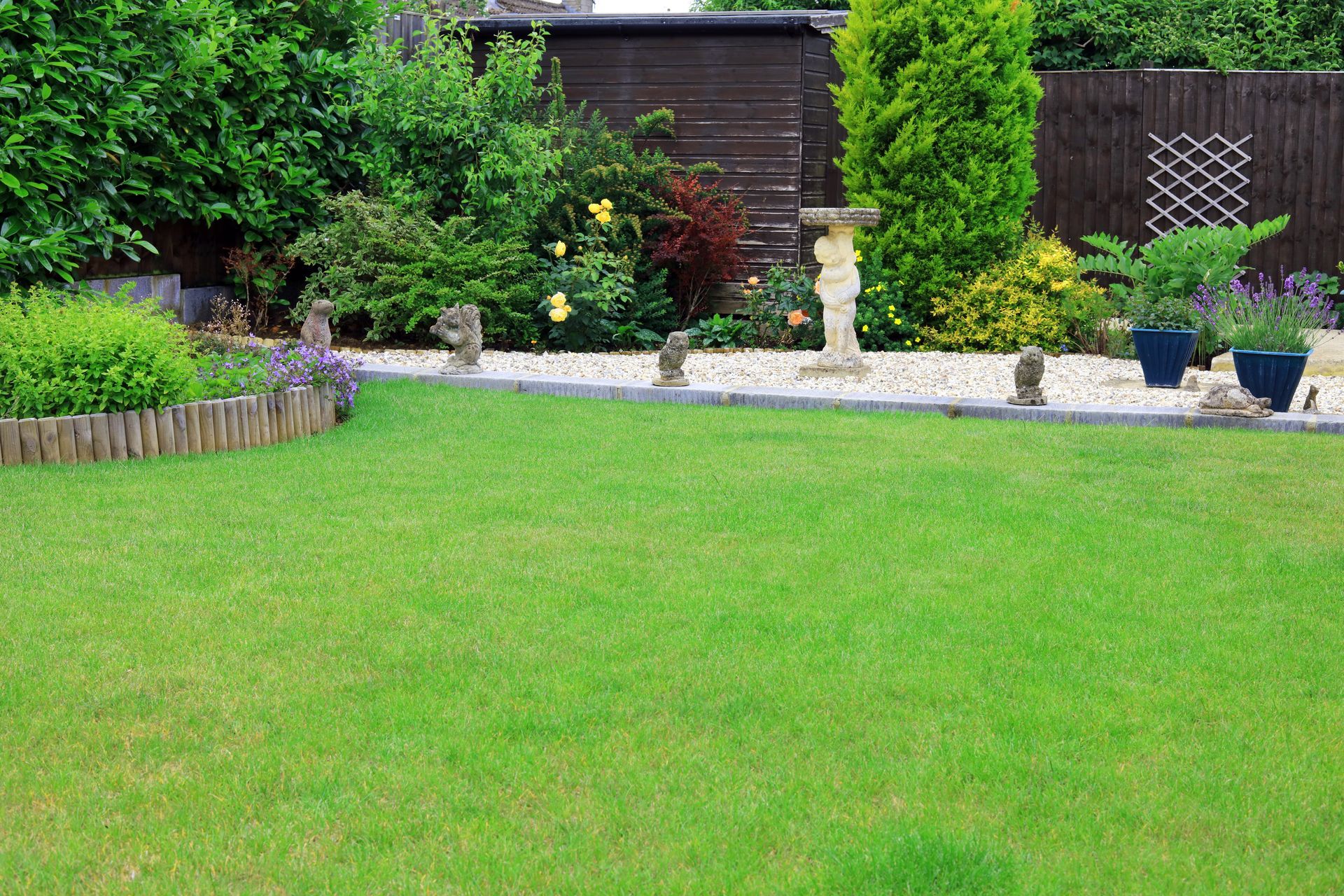 Pelouse verte bordée de gravier, agrémentée de statues et de plantes dans un jardin. Arbustes et clôture en arrière-plan.