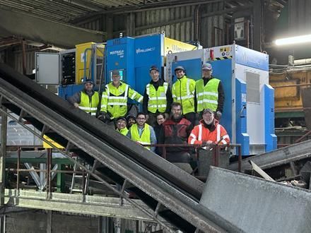 Eine Gruppe von Arbeitern in Sicherheitswesten steht vor Recyclingmaschinen in einer Anlage.