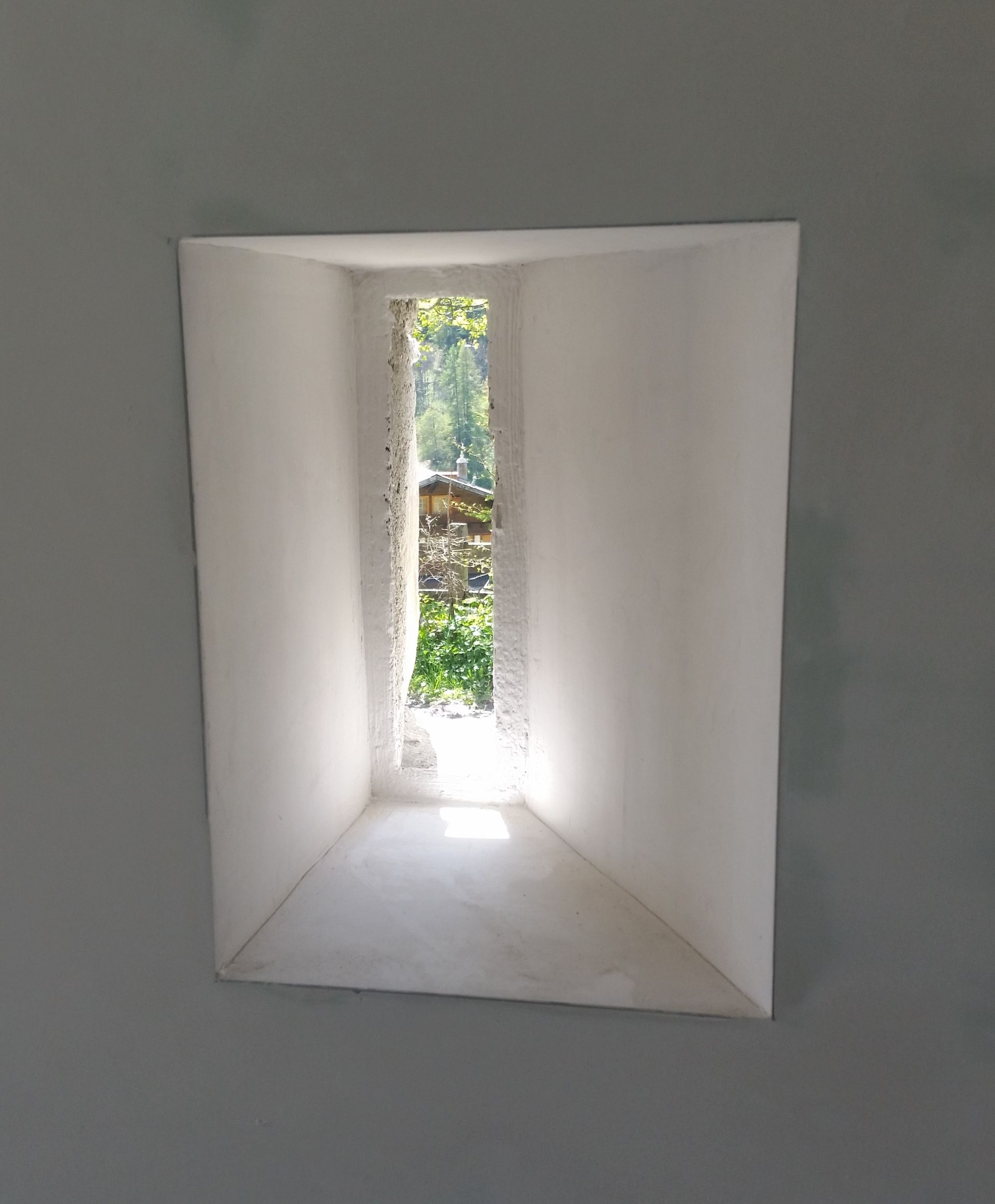 Une fenêtre carrée dans un mur blanc avec vue sur un jardin.