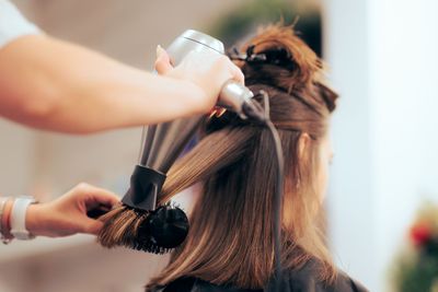 Femme aux longs cheveux bruns ondulés posant sur un fond beige, vêtue d'un haut noir à épaules dénudées
