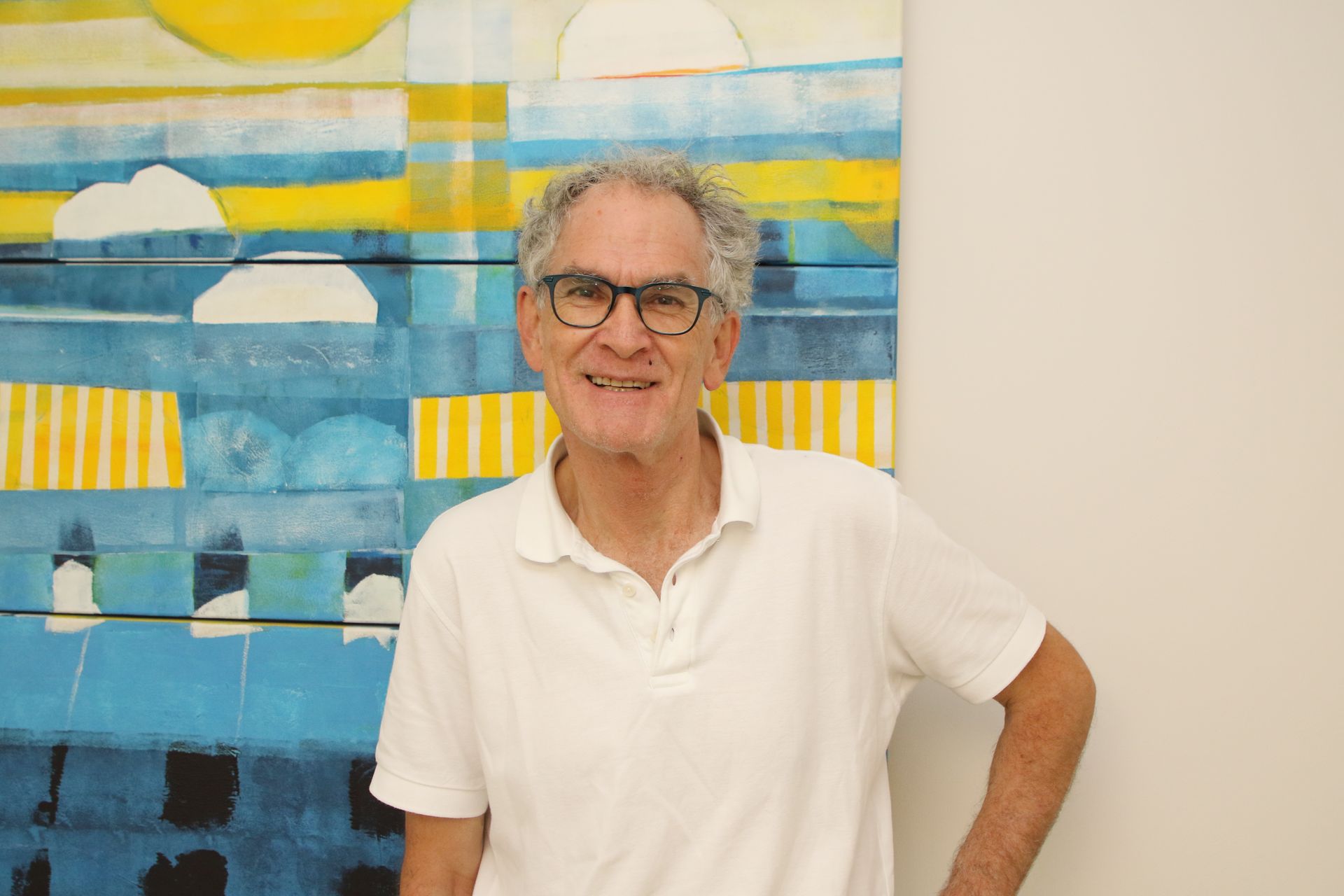 Mann mit Brille lächelt, steht neben einem blau-gelb-weißen abstrakten Gemälde. Weißes Hemd.