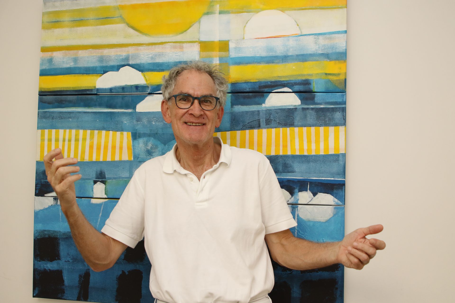 Mann steht mit ausgestreckten Armen vor einem abstrakten blau-gelb-weißen Gemälde und lächelt.