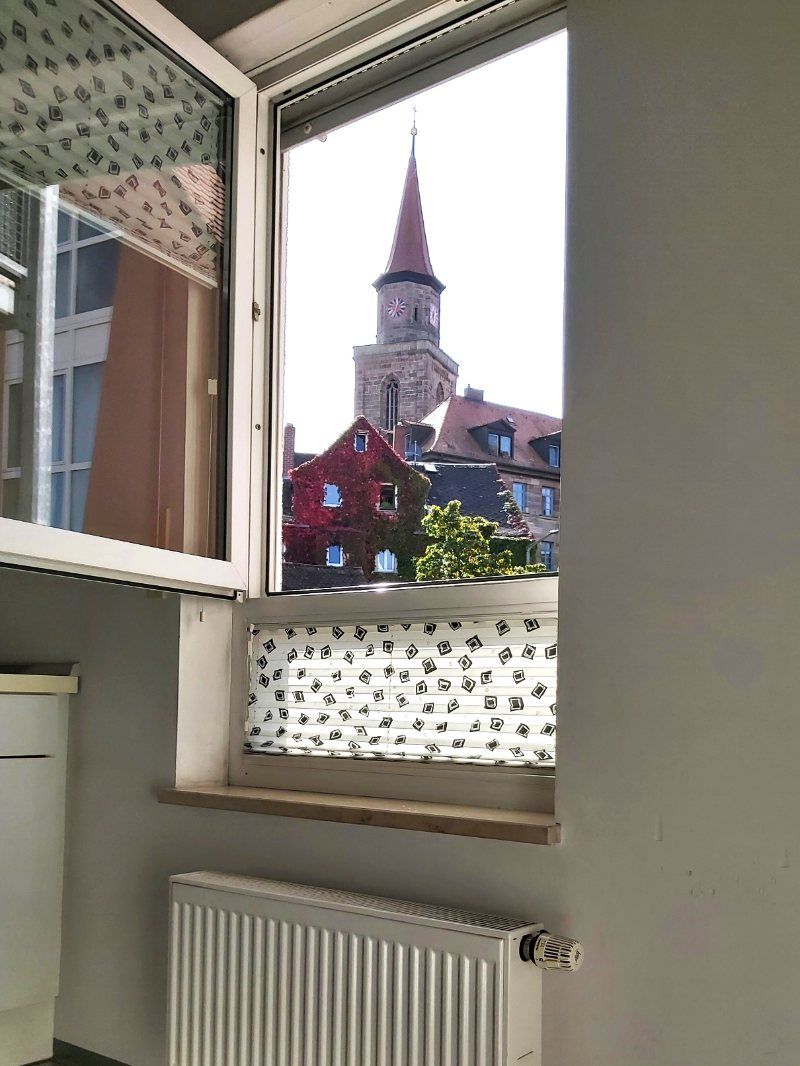 Offenes Fenster mit Blick auf einen Kirchturm und Gebäude mit roten Dächern. Unter dem Fenster befindet sich ein Heizkörper.