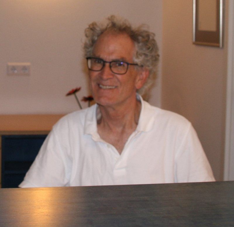 Mann mit lockigem grauem Haar und Brille lächelt. Trägt ein weißes Hemd. Drinnen, unscharfer Hintergrund.