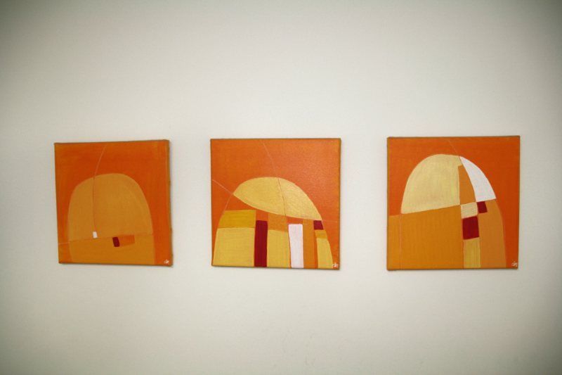 Drei quadratische abstrakte Gemälde mit orangefarbenen und goldenen Formen auf einer weißen Wand.