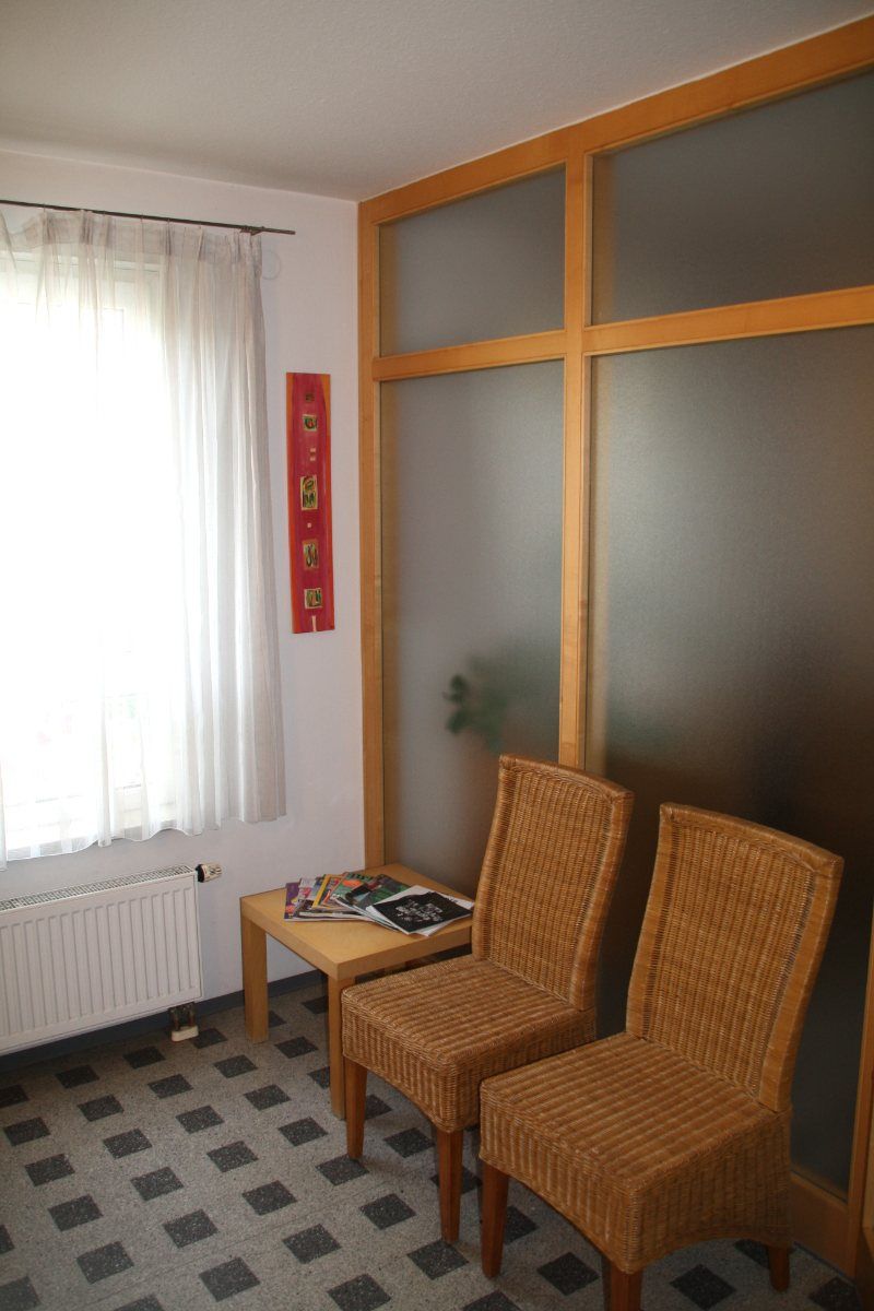 Wartezimmer mit zwei Korbstühlen, kleinem Tisch und Milchglastrennwand.