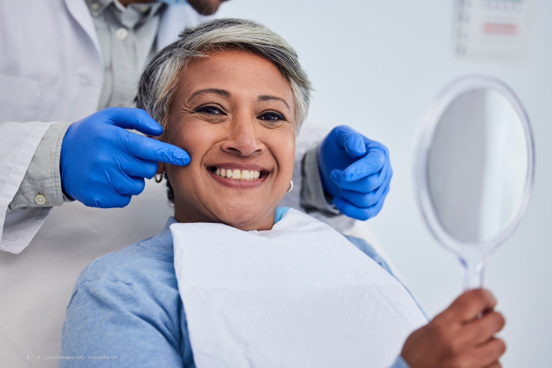 Frau in der Zahnarztpraxis lächelt, während sie in einen Handspiegel schaut. Zahnarzt in blauen Handschuhen zeigt auf ihre Wange.