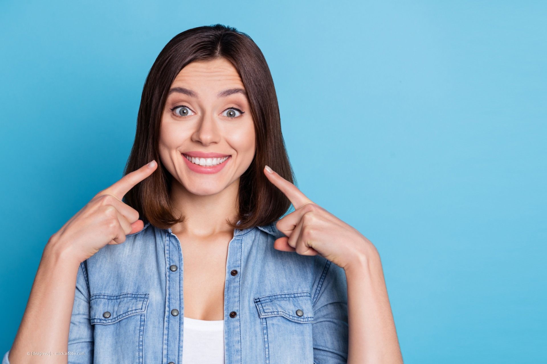 Frau zeigt auf ihr breites Lächeln, hellblauer Hintergrund.