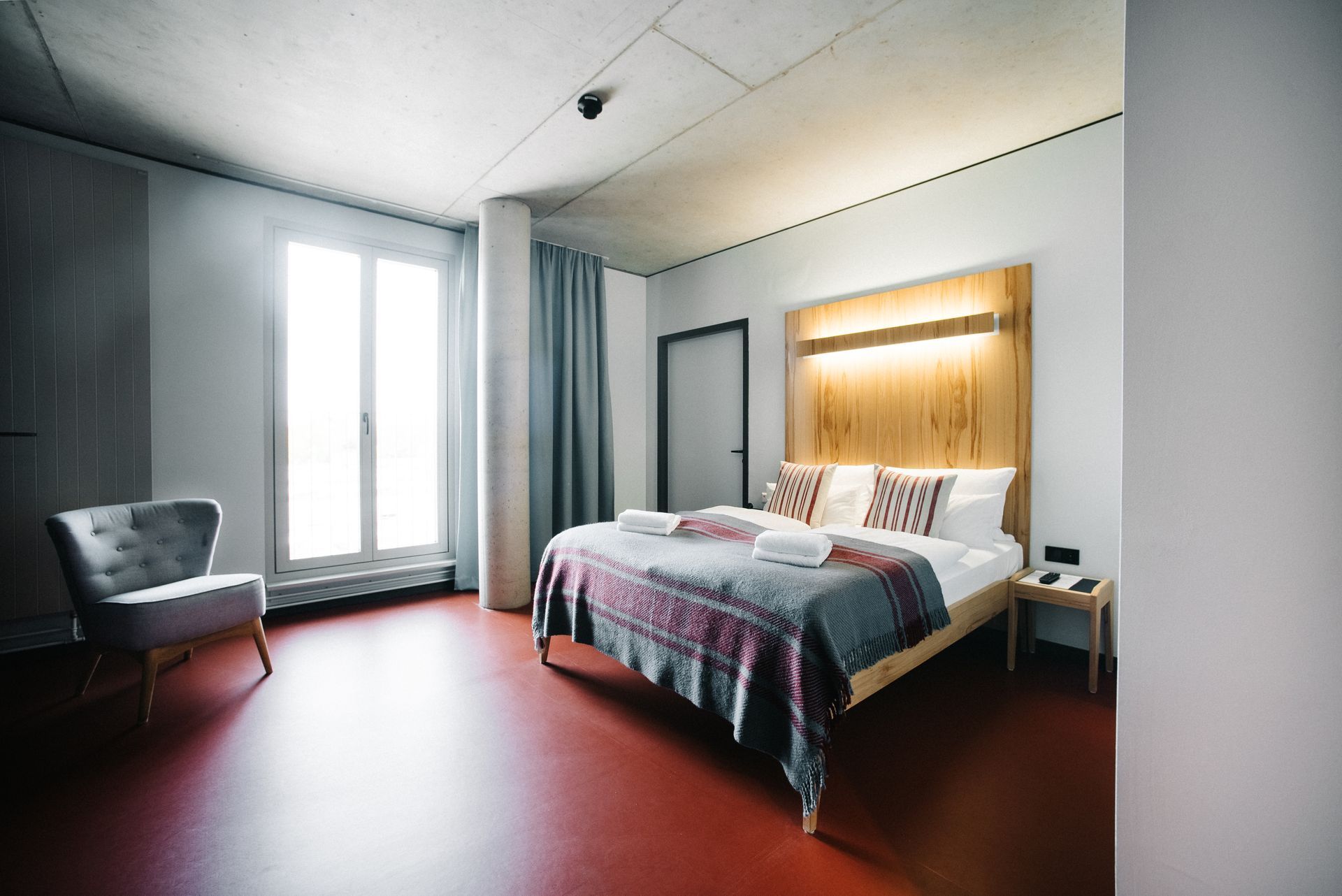 Schlafzimmer mit Bett, Stuhl und großem Fenster. Roter Boden, hölzernes Kopfteil und graue Vorhänge.