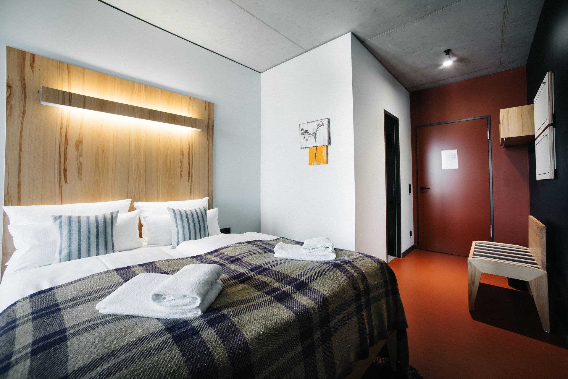 Hotelzimmer mit Bett, karierter Decke und weißen Handtüchern. Holzkopfteil und Akzentwand, rote Tür.