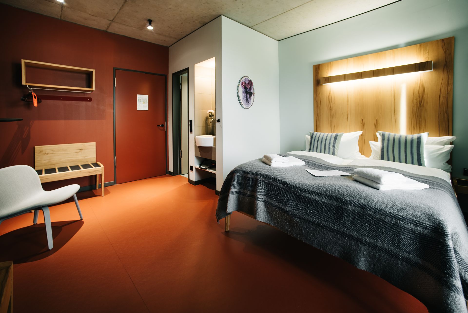 Hotelzimmer mit rotem Boden, Bett, hölzernem Kopfteil, Stuhl, Bank und roter Tür.