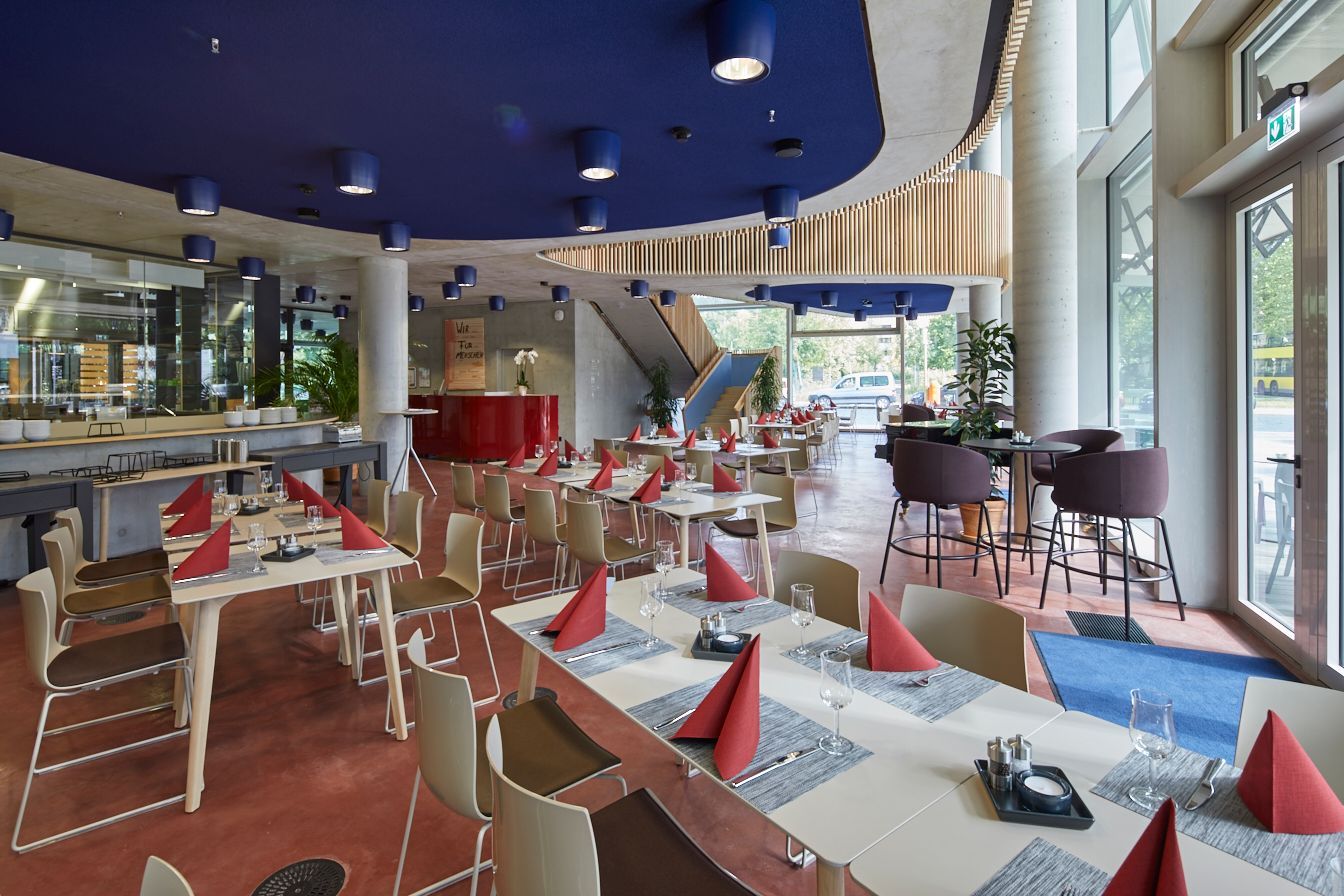 Restaurantinterieur mit gedeckten Tischen; rote Servietten und Tische, blaue Decke, Treppe im Hintergrund.