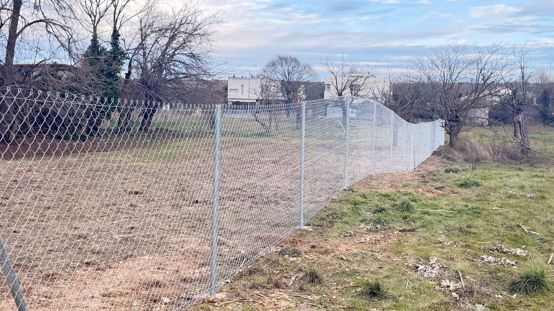 Valla de alambre en una zona cubierta de hierba, con árboles y edificios al fondo bajo un cielo nublado.