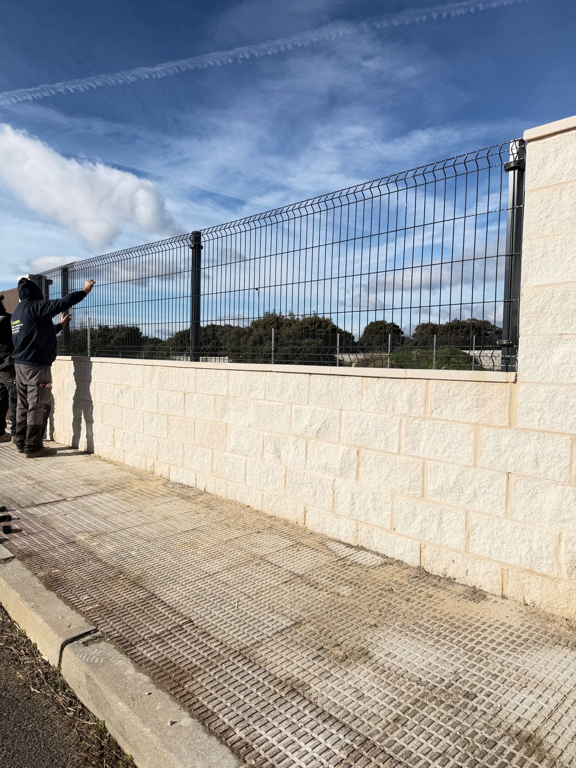 Una persona señala una valla metálica decorativa sobre un muro de piedra, contra un cielo azul.