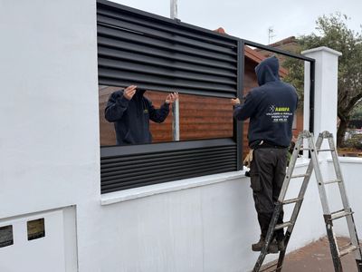 Dos trabajadores instalando una persiana negra en una pared blanca, utilizando una escalera en el exterior.