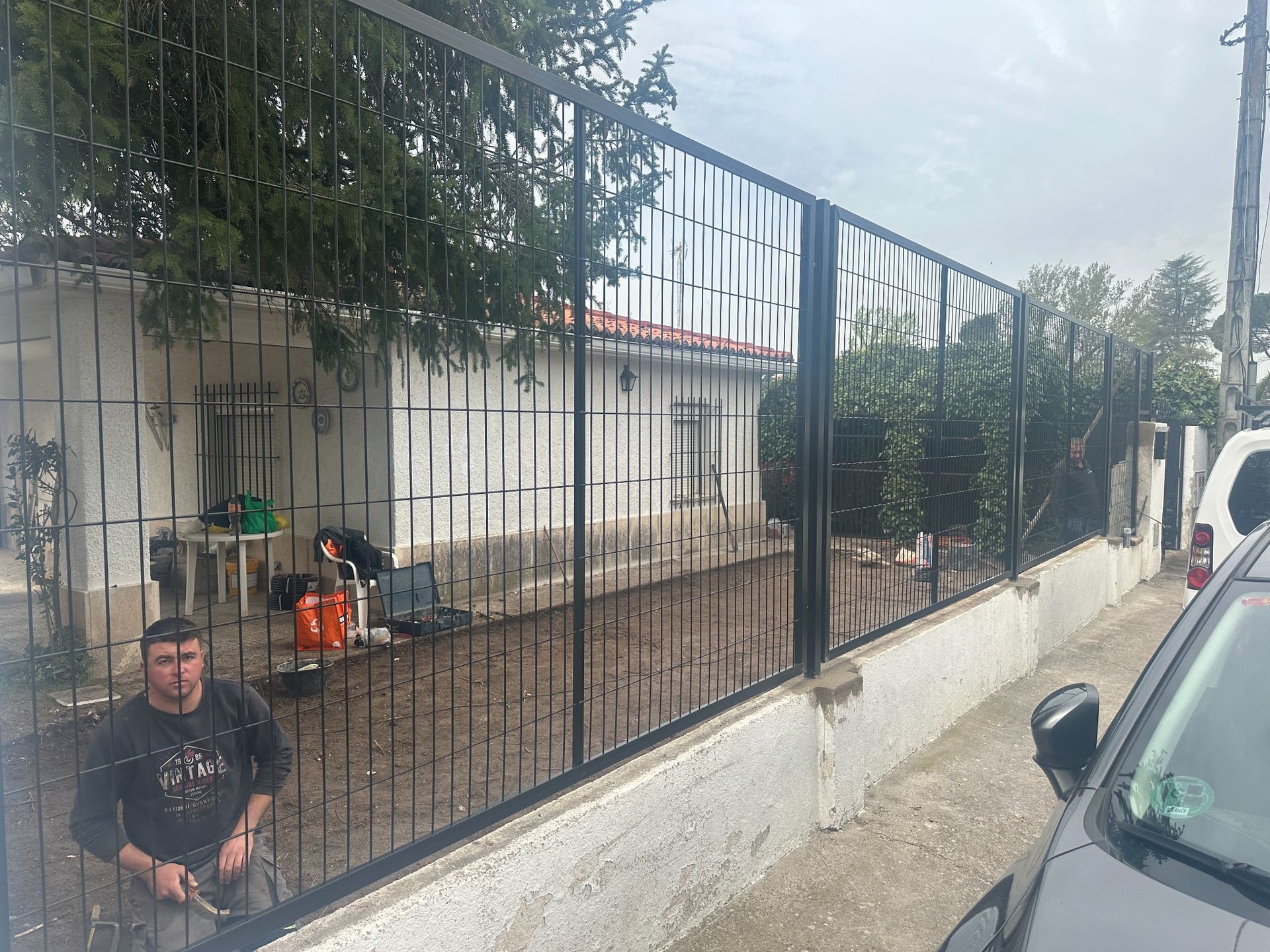 Hombre de pie junto a una valla metálica negra, al lado de un edificio y un coche aparcado.