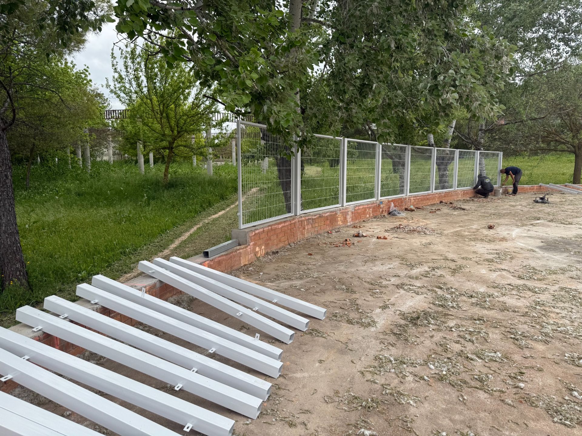 Construcción de una valla sobre una base de ladrillo curvado, con postes metálicos blancos y trabajadores en un entorno exterior con césped.