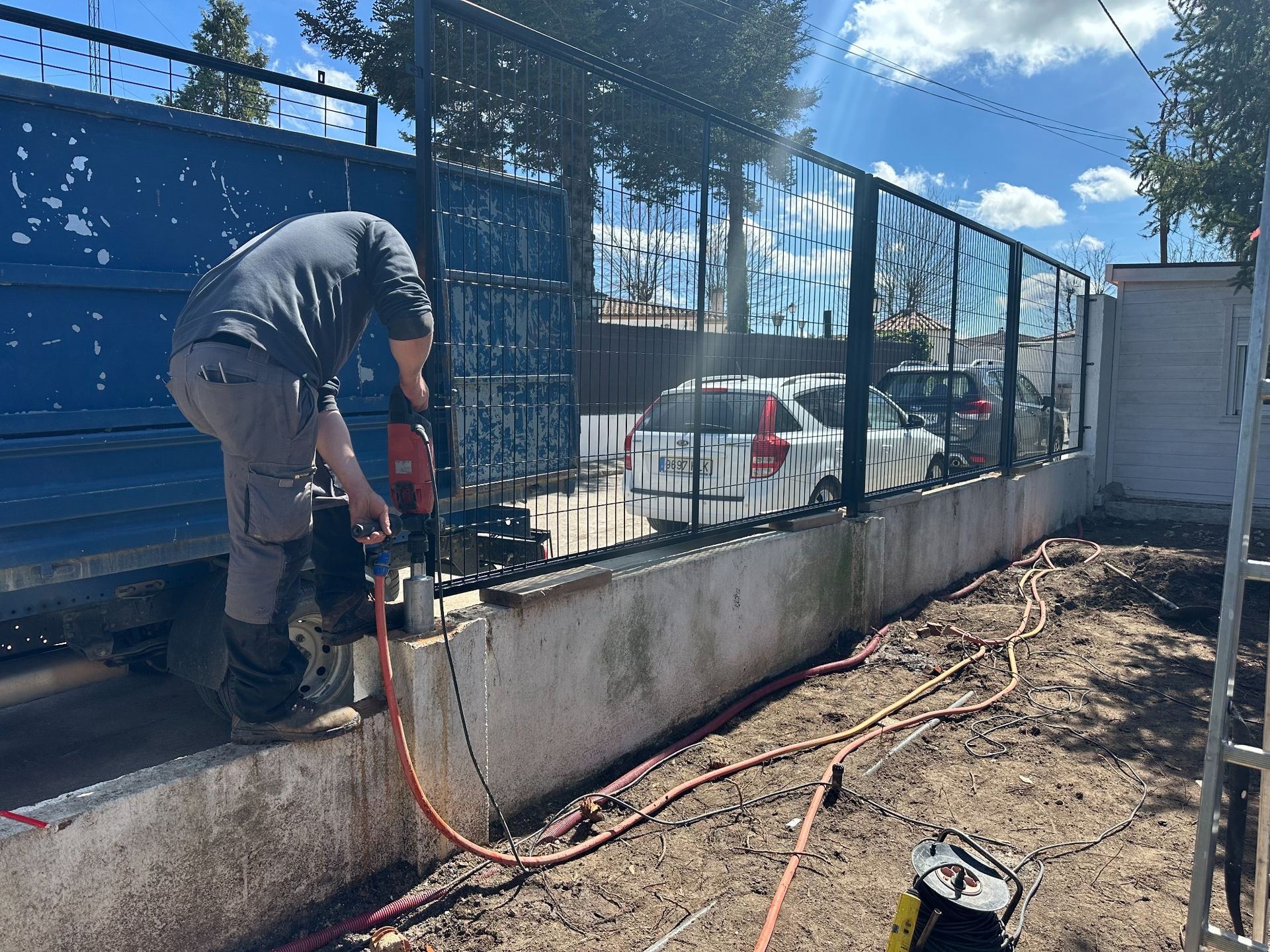 Persona cortando un muro de hormigón con una herramienta. Al fondo, una valla negra y un coche aparcado. Día soleado.