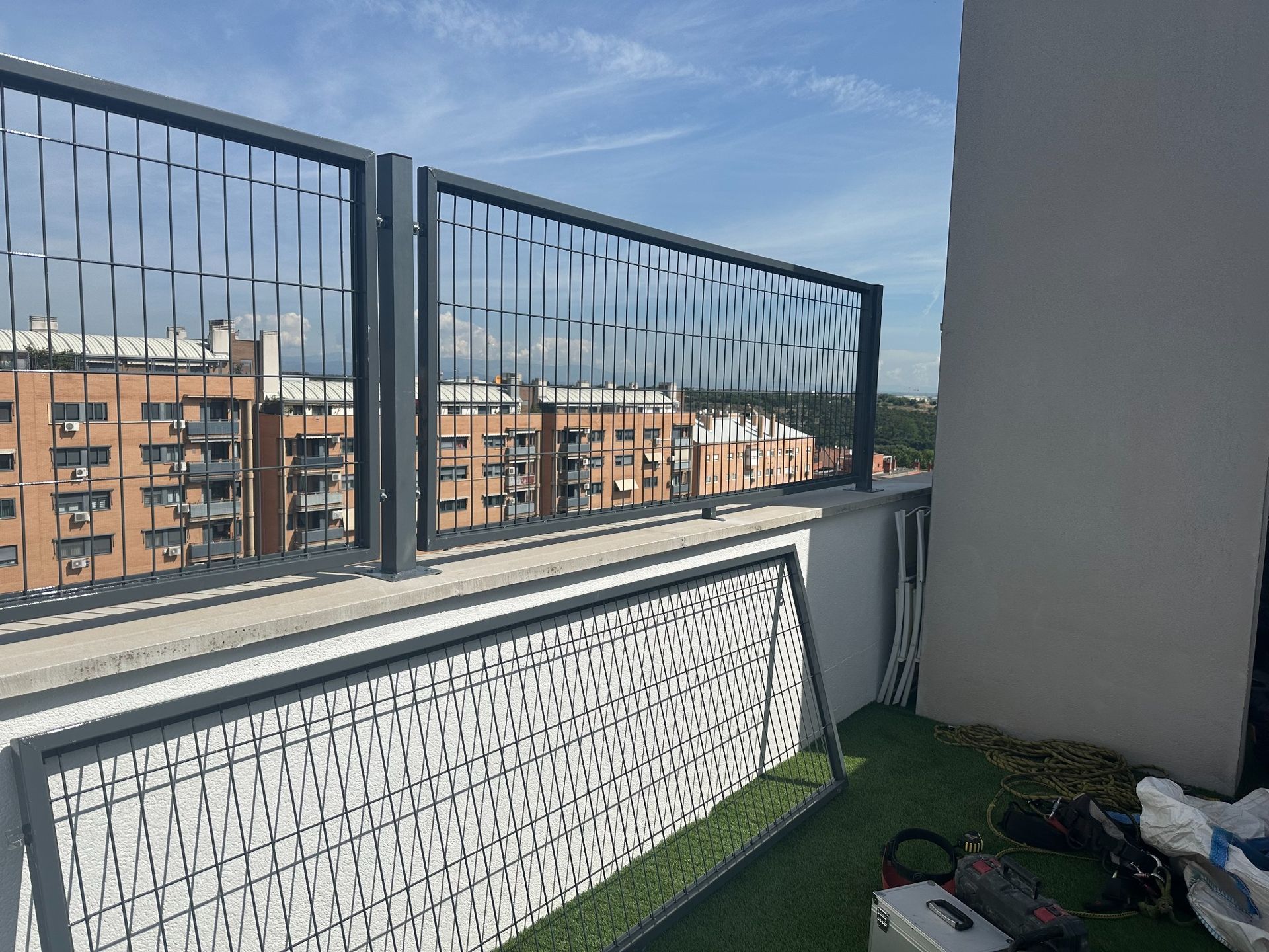 Secciones de valla metálica en un balcón con vistas a edificios urbanos bajo un cielo azul.