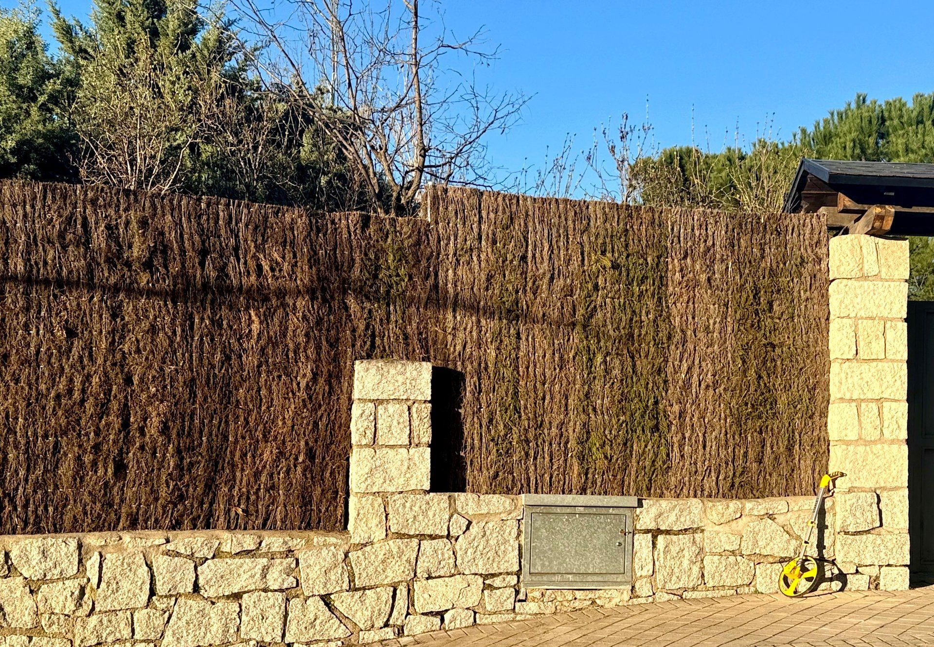 Muro de piedra rematado con una valla de bambú marrón. Un panel metálico está incrustado en la piedra.
