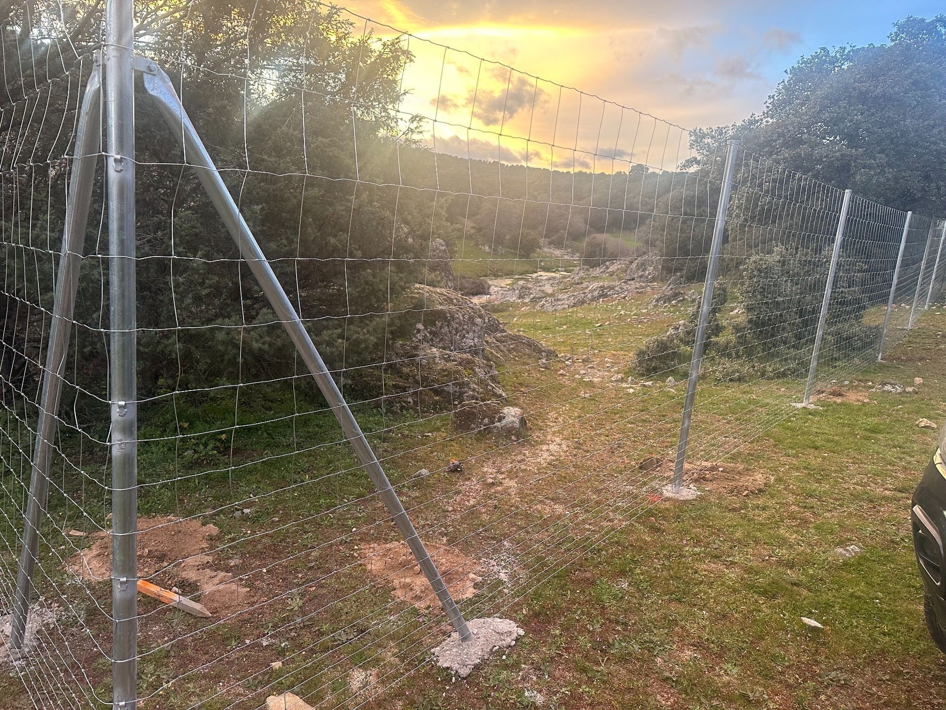 Valla galvanizada en un campo de hierba con una puesta de sol borrosa al fondo.