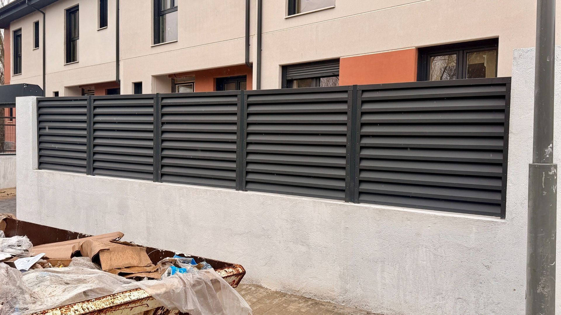 Valla de listones horizontales de color gris sobre un muro de hormigón frente a un edificio.