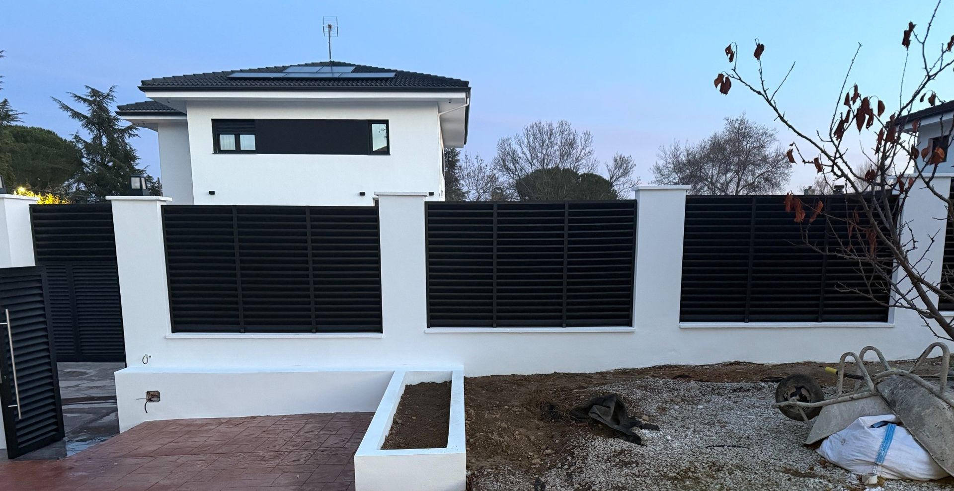 Casa blanca con valla y portón de listones negros; un pequeño jardín en el frente.