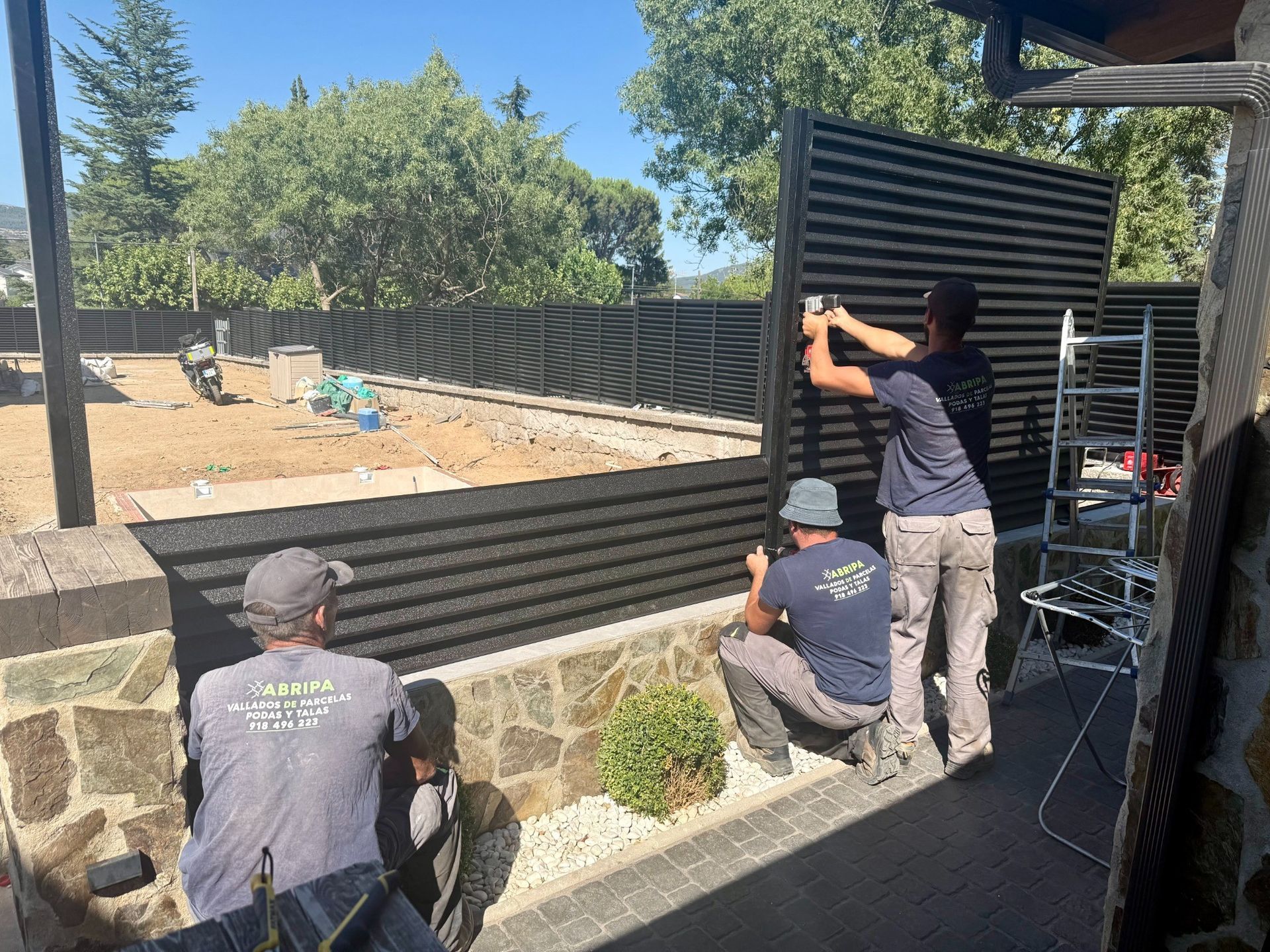 Tres trabajadores instalando una valla de listones negros en un patio, en un día soleado.