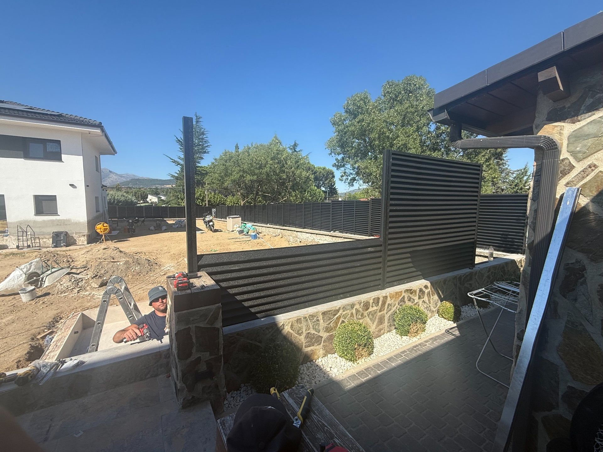 Obra con valla parcial, mampostería y un hombre trabajando. Día soleado.