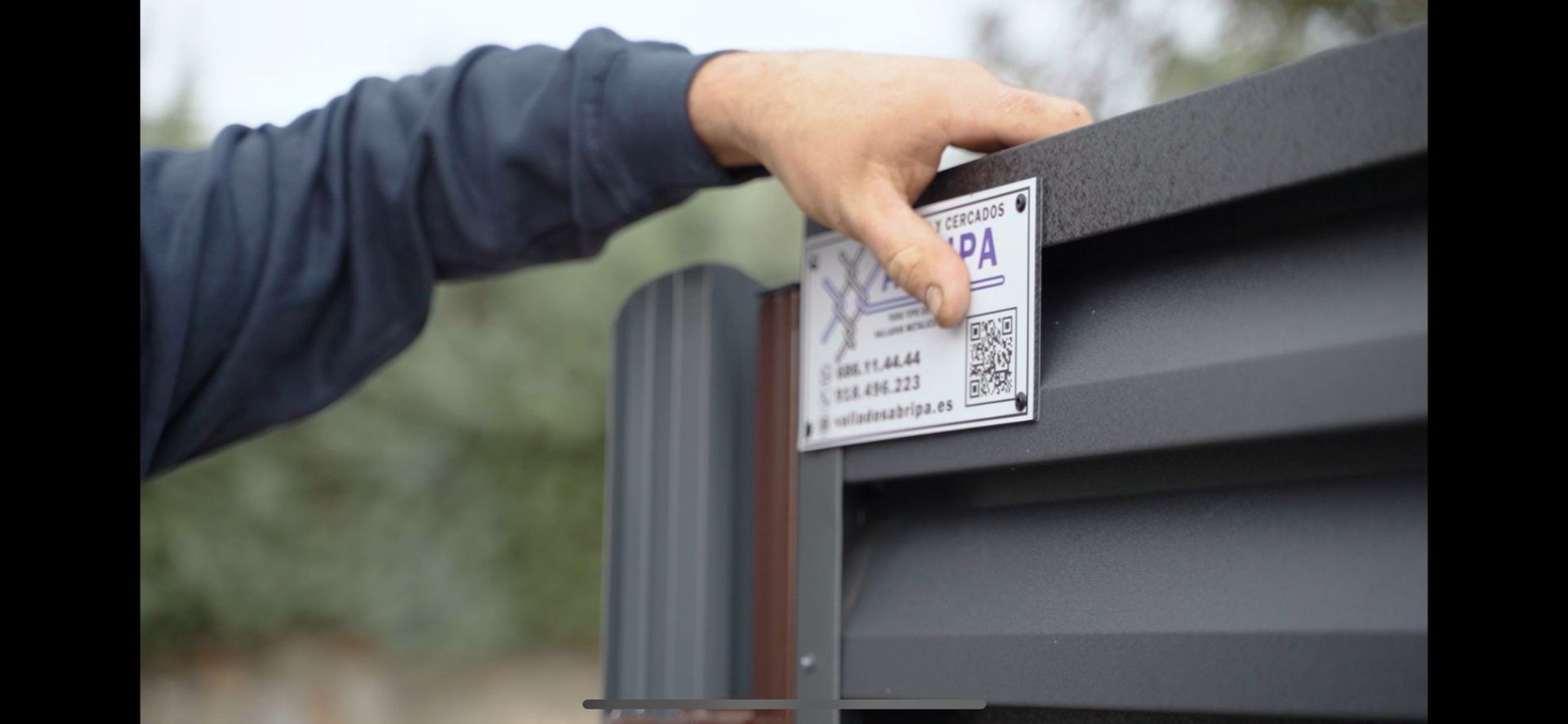 Mano de una persona sobre una valla metálica con una etiqueta blanca. La etiqueta incluye texto, un código QR y el logotipo de la empresa.