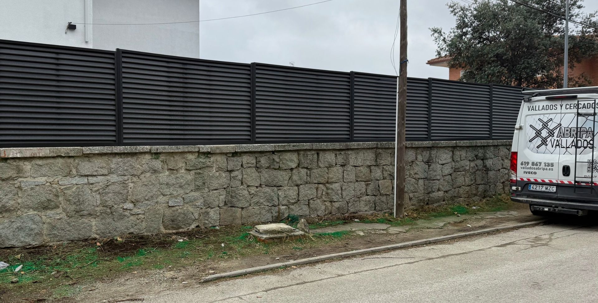Una valla oscura de listones horizontales sobre un muro de piedra. Una furgoneta está estacionada a la derecha.