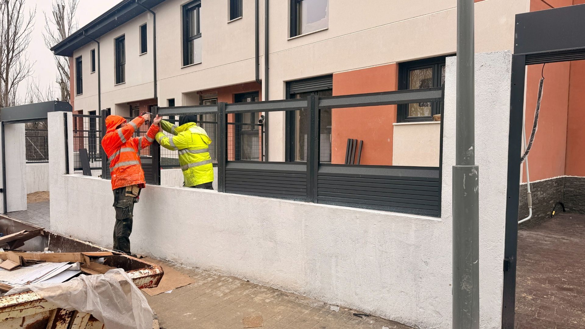 Obreros de la construcción instalan una valla metálica oscura sobre una pared blanca cerca de un edificio.