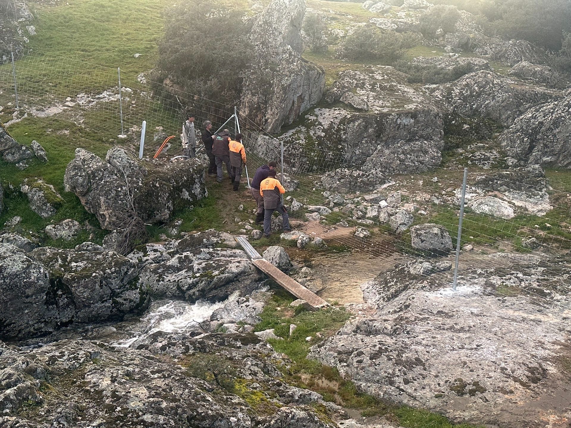 Personas vestidas con uniforme de trabajo de color naranja en un terreno rocoso cerca de un pequeño puente de madera.