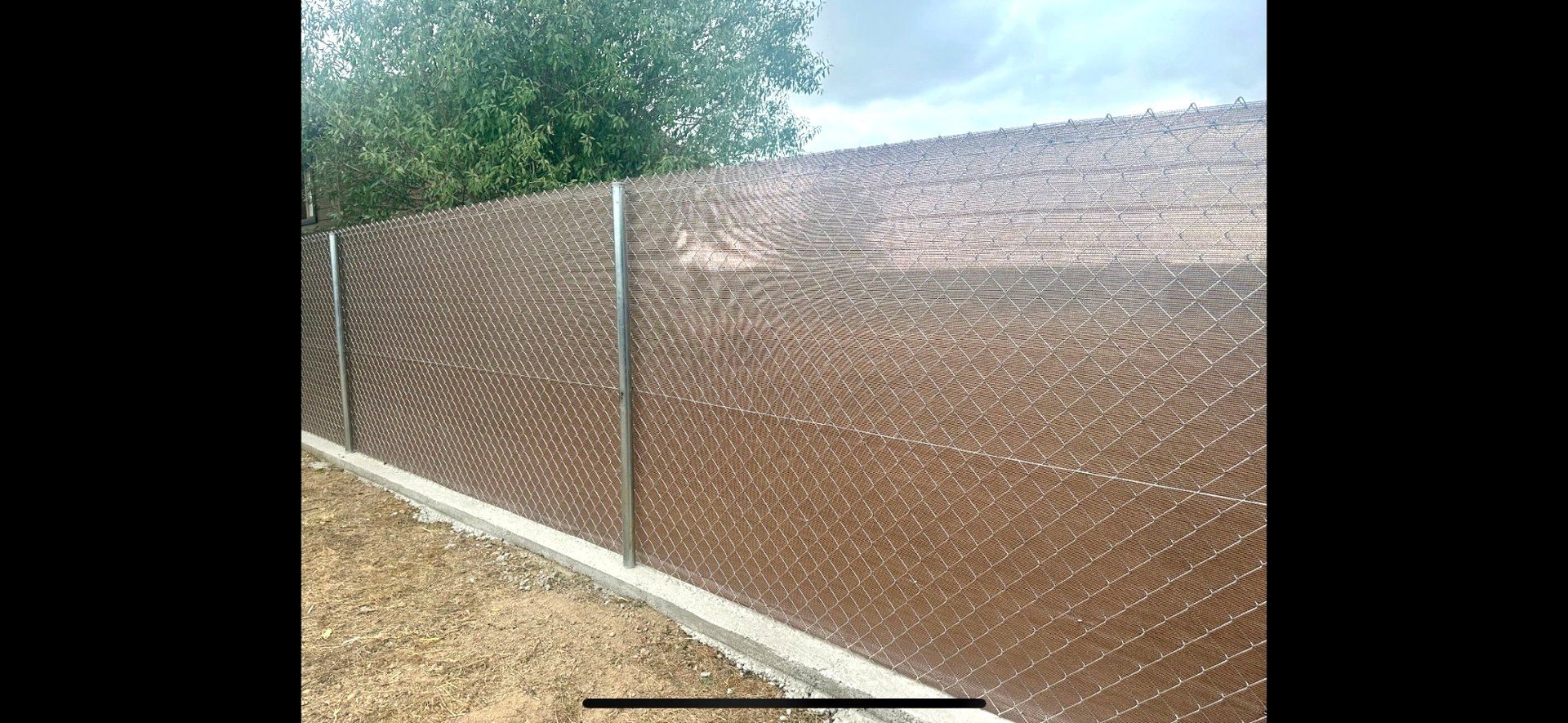 Una cerca de alambre marrón con postes metálicos. Un borde de hormigón recorre la parte inferior. Árboles verdes al fondo.