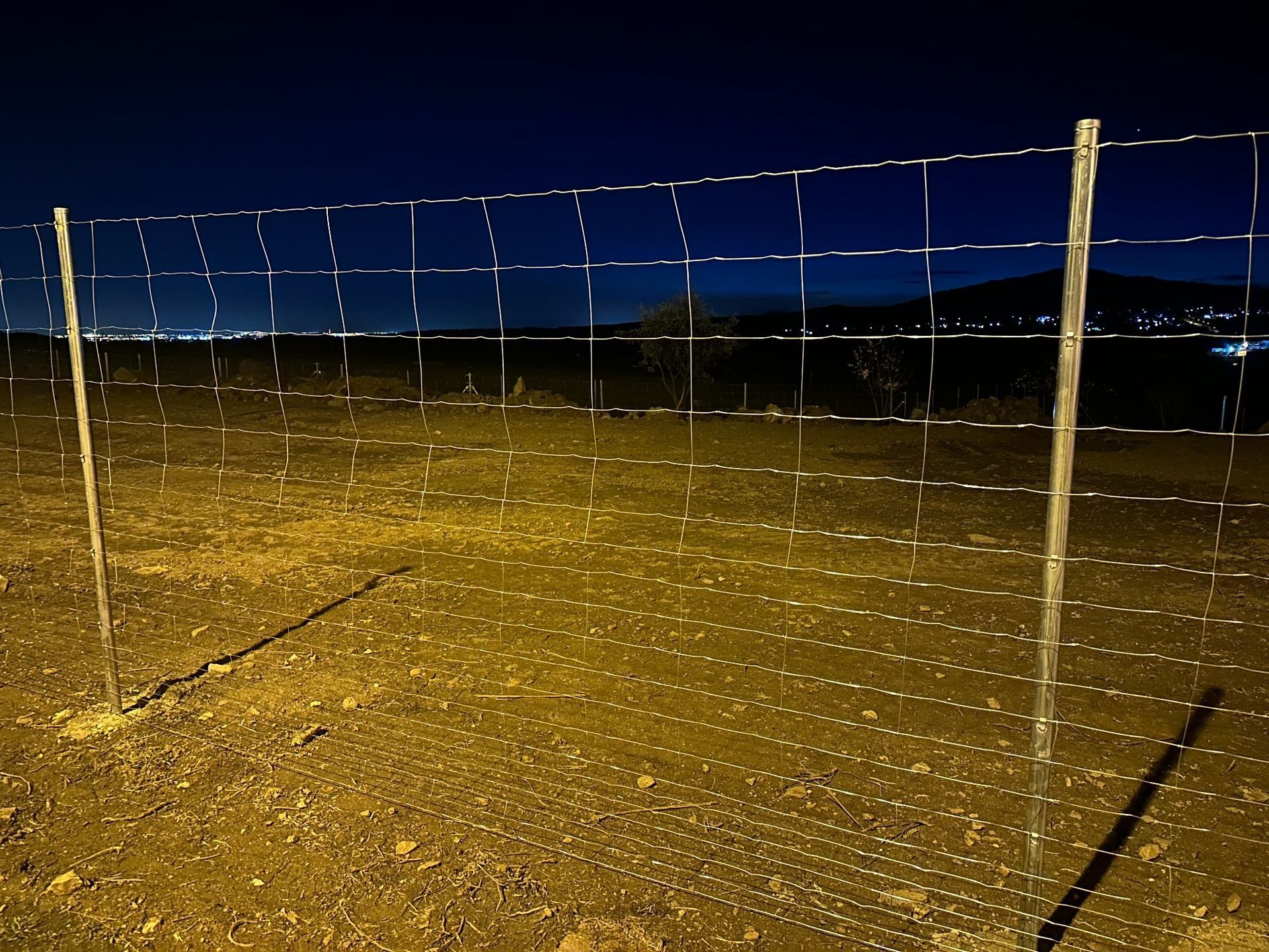 Una cerca de alambre en un campo de tierra por la noche, luces de la ciudad visibles en la distancia.