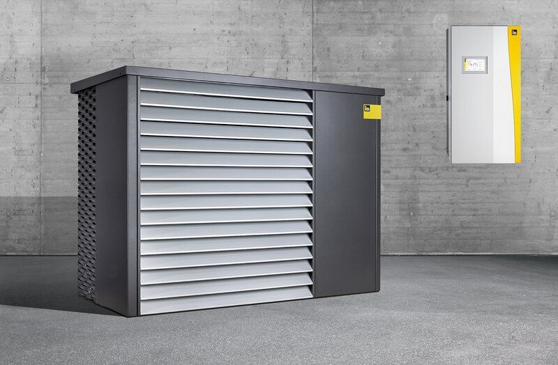 Graue und schwarze Maschinen mit Lamellenverkleidung, daneben ein weißer Kasten mit gelben Akzenten an einer Betonwand.