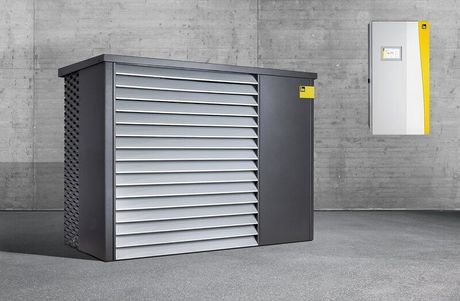 Graue und schwarze Maschinen mit Lamellenverkleidung, daneben ein weißer Kasten mit gelben Akzenten an einer Betonwand.